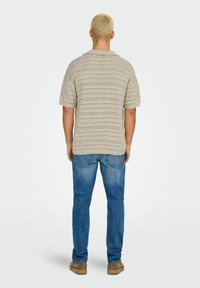Crème gekleurde, korte mouwen gebreide shirt met open patroon, gedragen met blauwe spijkerbroek en bruine schoenen, gezien vanuit de achterkant.