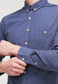 Modrá košile na knoflíky z texturované látky, s malou náprsní kapsou, dřevěnými knoflíky a klasickým límcem.