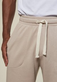 Mann trägt beigefarbene Sweathosen mit Zugband, hat die Hand in der linken Tasche und ein weißes T-Shirt, das reingesteckt ist.