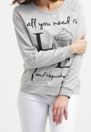 Sudadera gris de mangas largas, con el texto negro "todo lo que necesitas son L y cupcakes" y un gráfico de cupcake. Pantalones blancos.
