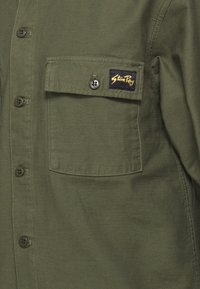 Green button-up shirt with a chest pocket. Features a stitched logo tag and matching buttons. Material appears soft and durable.
