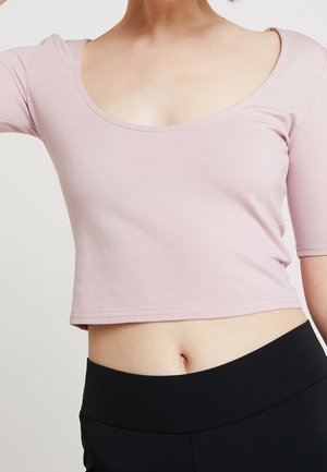 Nainen, joka käyttää vaaleanpunaista lyhytmallista toppia, jossa on kolme neljäsosaa hiha ja korkeavyötäröiset mustat housut, paljastaen vyötärön ja navan.