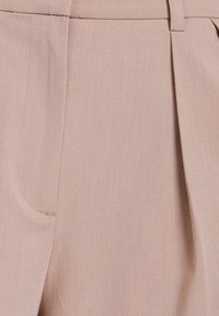 Beige skräddarsydda byxor med en slät textur, med framfickor och en veckad design. Tyget visar ett subtilt snedrutigt mönster.