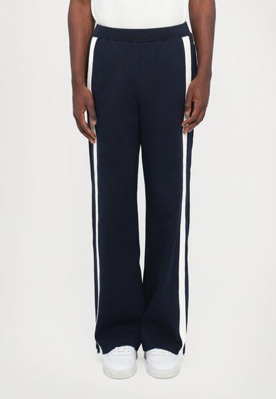 Pantalons de sport navy avec des rayures blanches sur les côtés, fabriqués en tissu doux, présentant une coupe décontractée et une taille élastique. Portés avec des baskets blanches.