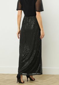 Gonna maxi nera con paillettes scintillanti, tessuto morbido e vita alta. Abbinata a una blusa nera con maniche trasparenti e tacchi alti.