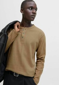Joven con una camiseta henley de manga larga marrón y pantalones negros, sosteniendo una chaqueta negra sobre el hombro, mirando hacia un lado.