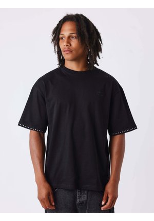 Jeune homme avec des dreadlocks de longueur moyenne portant un t-shirt noir oversize avec des bords de manches cloutés, regardant sur le côté sur fond blanc.