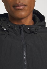 Gros plan d’une personne portant une veste à capuche noire avec une fermeture éclair à l’avant et des cordons réglables sur la capuche.