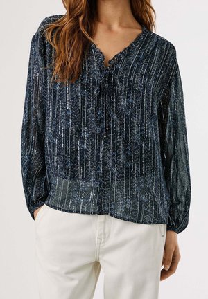 Femme portant un chemisier à manches longues noir et bleu à motifs avec un lien au niveau de l'encolure, associé à un pantalon écru.