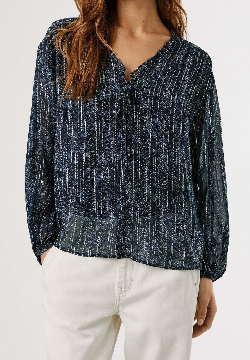 Femme portant un chemisier à manches longues noir et bleu à motifs avec un lien au niveau de l'encolure, associé à un pantalon écru.