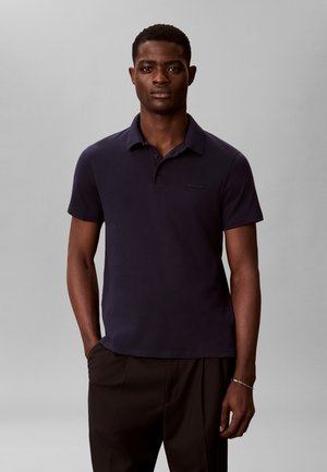 Młody mężczyzna stojący, ubrany w granatową koszulkę polo z krótkim rękawem i czarne spodnie, na jednolitym jasno-szarym tle.