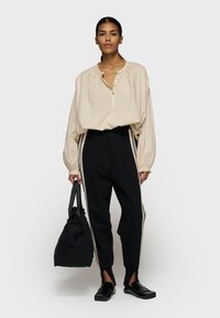 Donna che indossa una blusa beige con bottoni, pantaloni sportivi neri con strisce laterali, mocassini neri, tiene una borsa a mano nera, in piedi su uno sfondo bianco.