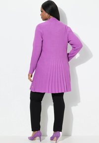 Lila gerippter Cardigan mit einem plissierten Rücken design, kombiniert mit schwarzen Hosen und lila Wildlederschuhen mit hohen Absätzen. Glatte Textur und taillierte Passform.