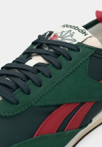 Sapatilha verde e azul escuro com riscas laterais vermelhas, atacadores pretos e uma língua branca com o logotipo da Reebok e um design de estrela.