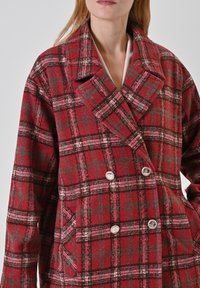 Donna che indossa un cappotto doppiopetto a quadri rossi e neri con grande colletto e quattro bottoni argento, mani nelle tasche, contro uno sfondo semplice.