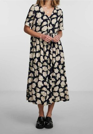 Kvinde iført en sort midi-kjole med hvidt blomsterprint, korte ærmer, indsnøret talje, lagdelte halskæder og sorte loafers, stående med hænderne foldet.