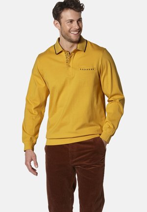 Long-sleeve polo shirt in mustard yellow cotton, featuring a patterned collar, button placket, and a front pocket with horizontal slits. Brown corduroy pants.