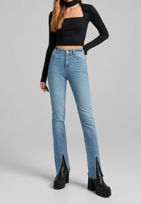 Hoch taillierte hellblaue Jeans mit fransigem Saum und seitlichen Schlitzen, kombiniert mit einem figurbetonten schwarzen Langarmshirt und klobigen schwarzen Plateaustiefeln.