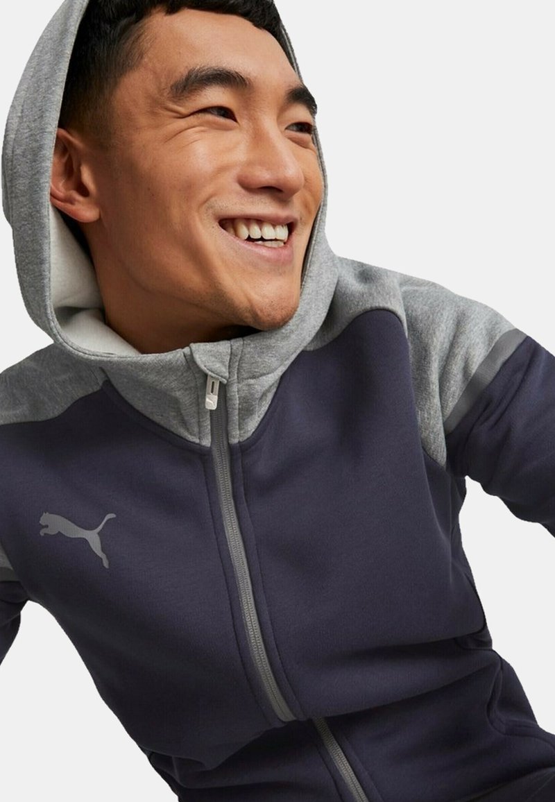 Felpa con zip di colore navy con accenti grigi, dotata di zip frontale, cappuccio con cordino e logo riflettente sul petto. Tessuto morbido.