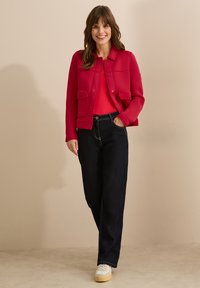 Röd, kort jacka med två framfickor, lager på en röd topp, i kombination med mörka jeans och vita sneakers. Enkel, avslappnad design.