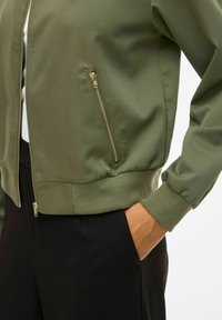 Olivgrüne Bomberjacke mit gerippten Bündchen und Saum, ausgestattet mit goldenen Reißverschlüssen an den Seitentaschen und einer glatten, matten Textur.