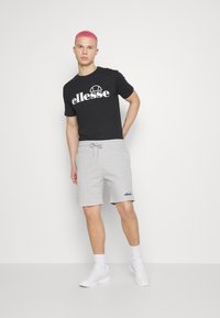 Čierne tričko s veľkým bielym logom "ellesse", v kombinácii so sivými šortkami s malým čiernym logom a bielymi teniskami s členkovými ponožkami.