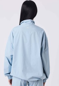 Giacca oversize azzurro chiaro con collo alto, polsini elastici e un piccolo dettaglio a stella sulla schiena. Realizzata in tessuto liscio e leggero.