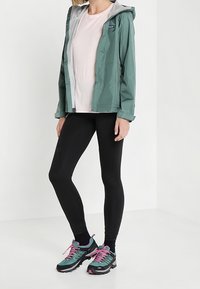 Letvægts grøn regnjakke med grå foring, parret med sorte leggings og teal sneakers med pink accenter. Simpelt og funktionelt design.