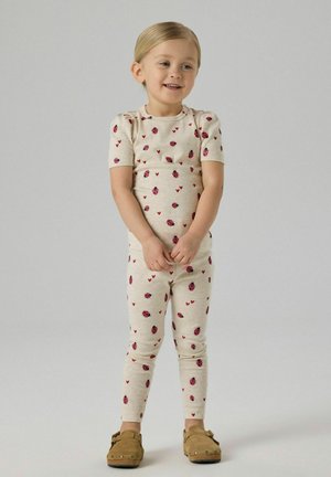 Junges Kind steht, trägt beige Outfit mit roten Marienkäfern und Herzmustern sowie braune Slipper, lächelt leicht, heller Hintergrund.