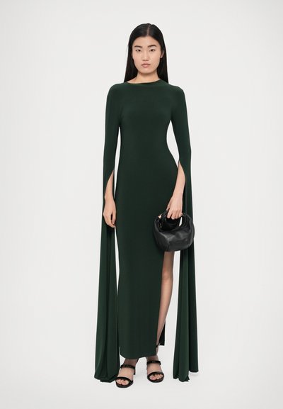 Zöld hosszú ujjú ruha oldalsó hasítékokkal és lebegő anyagból. A modell egy fekete táskát tart és fekete, pántos szandált visel.