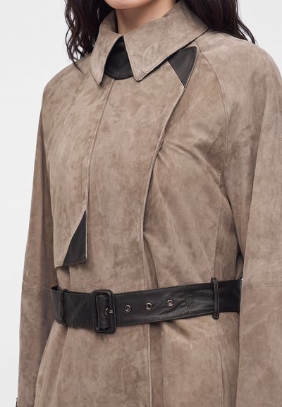 Trench-coat beige en daim avec large col, accents en cuir noir sur le col et la ceinture, porté par une personne aux cheveux foncés.