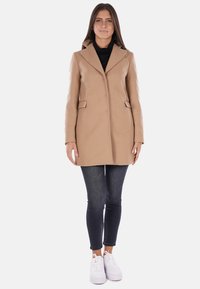 Cappotto in misto lana beige con una vestibilità slim, rever a falda, due tasche frontali e chiusura a bottone, abbinato a jeans scuri e sneakers bianche.