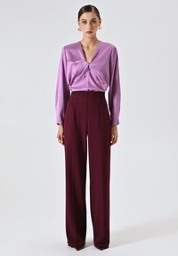 Blusa di satin lilla con scollo a V e dettagli arricciati nella parte anteriore, abbinata a pantaloni bordeaux a vita alta, strutturati con gamba dritta.