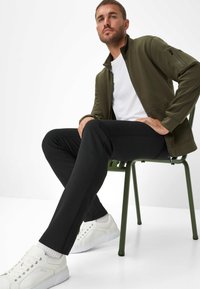 Giacca zip in verde oliva sopra una t-shirt bianca, abbinata a pantaloni neri e sneakers bianche. Il tessuto sembra liscio e informale.