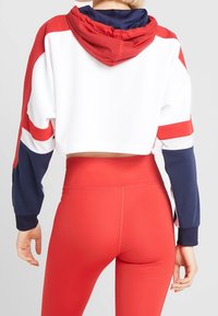 Donna che indossa una felpa corta bianca, rossa e blu navy con cappuccio rosso e leggings rossi a vita alta, vista da dietro.