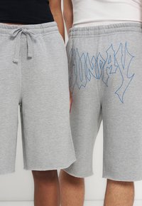 Dve osebi nosita svetlo sive flis kratke hlače, ene preproste z vrvico, druge z modrim grafičnim napisom "SUNDAY" na sprednji nogi.