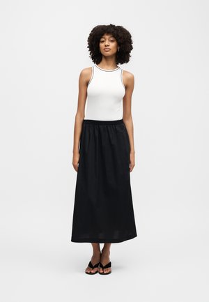 Γυναίκα με σγουρά μαλλιά στέκεται φορώντας λευκό αμάνικο τοπ, μαύρη μακριά φούστα και μαύρα σανδάλια με τακούνια σε απλό ανοιχτό φόντο.