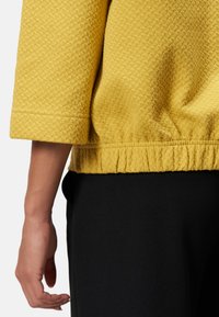 Sweatshirt jaune texturé avec des manches trois-quarts et un ourlet froncé, associé à un pantalon noir. Gros plan sur le tissu et le détail de la main.