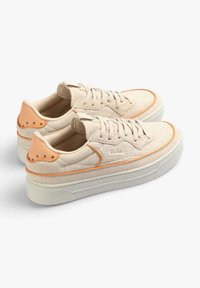 Baskets en toile beige avec dessus texturé, accents en cuir orange, bout rond et semelle à plateforme blanche. Présente un logo sur le côté et des clous décoratifs.