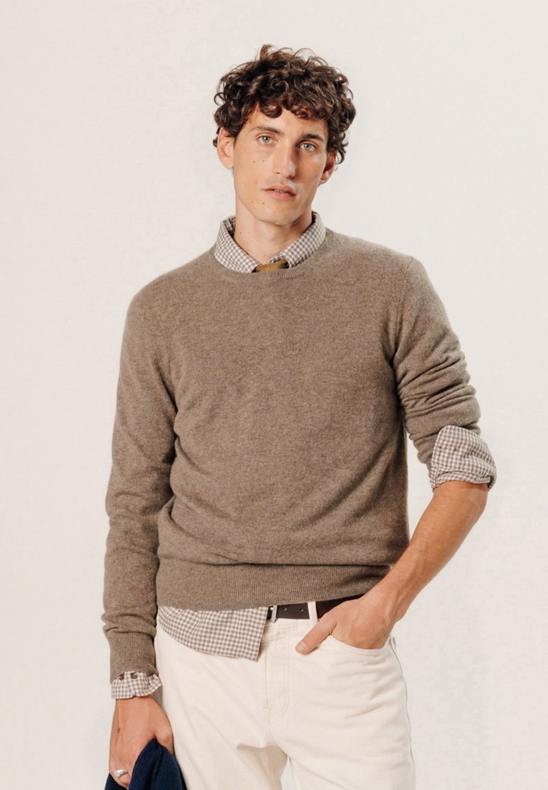 Brauner Strickpullover mit Rundhalsausschnitt über einem karierten Hemd mit Knöpfen. Das Model trägt weiße Hosen und hält eine blaue Jacke, was eine lässige Schichtung zeigt.