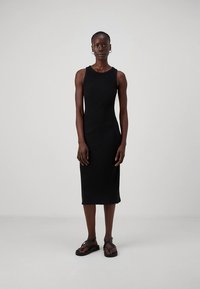 Černé žebrované midi šaty bez rukávů s přiléhavým střihem, hladkou texturou a jemnými ševními detaily. Nošeny s černými sandály.