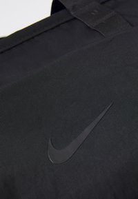 Fekete nylon bevásárlótáska kiemelkedő fekete Nike logóval. A textúra simának tűnik, finom fényléssel. Masszív fogantyúk láthatók.