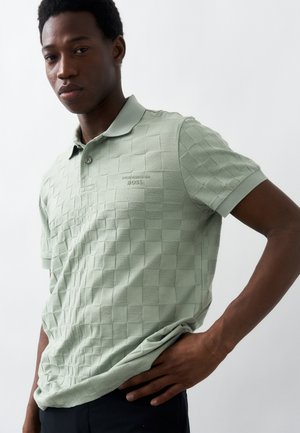 Νεαρός άνδρας που φοράει ανοιχτό πράσινο καρό πόλο με το λογότυπο "Porsche BOSS", στέκεται με το ένα χέρι στη μέση μπροστά σε λευκό φόντο.