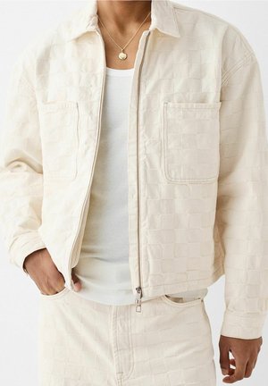 Mann trägt eine cremefarbene, kariert Jacke mit Fronttaschen, ein weißes, geripptes Tanktop, passende cremefarbene Hose und eine goldene Anhängerkette.