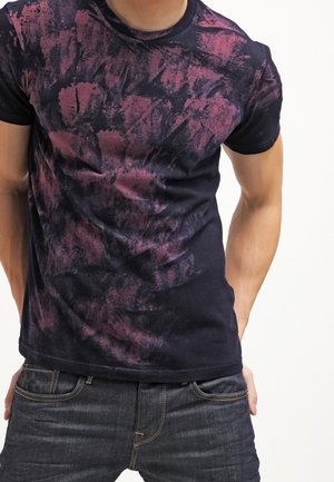 Mężczyzna noszący dopasowaną czarno-fioletową koszulkę tie-dye i ciemnoszare dżinsy, z rękami spoczywającymi blisko kieszeni.