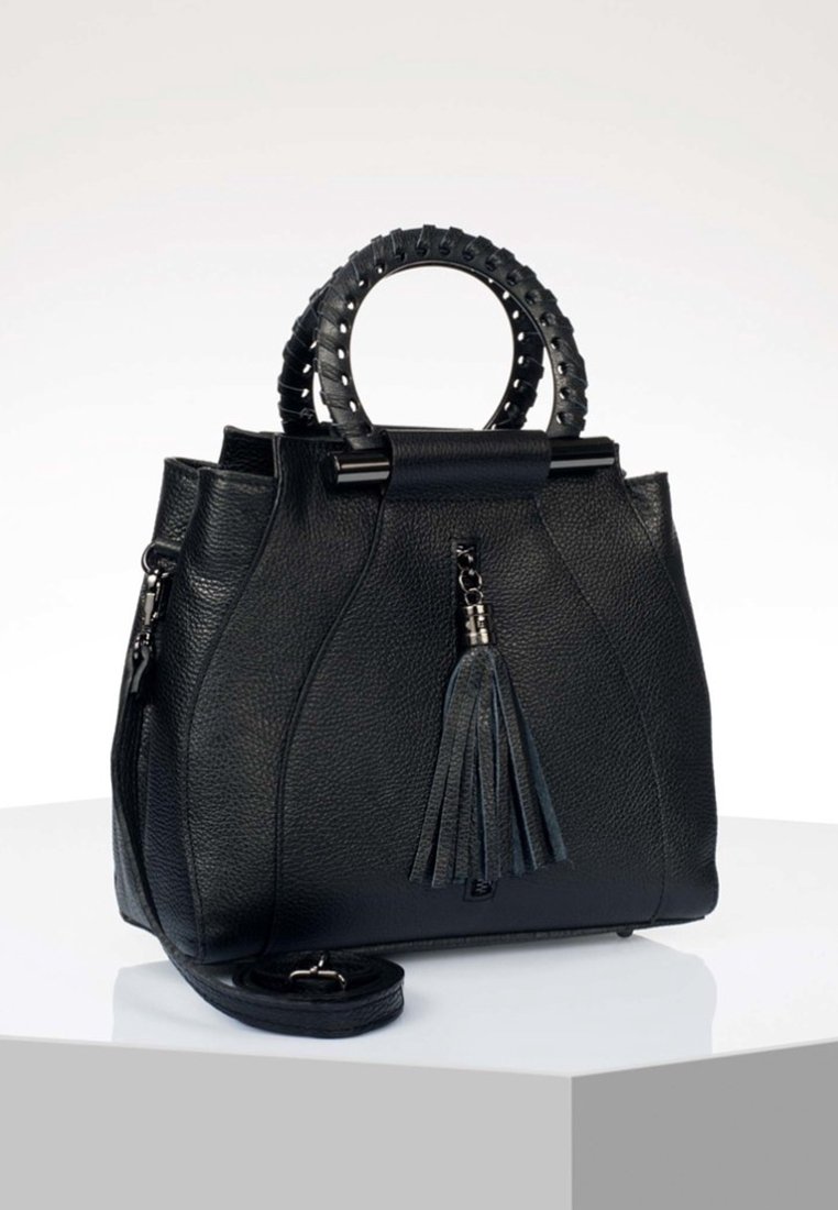 Sac à main en cuir noir texturé avec poignées circulaires enveloppées, détail de pampille à l'avant, fermeture éclair sur le côté et bandoulière amovible sur surface blanche.