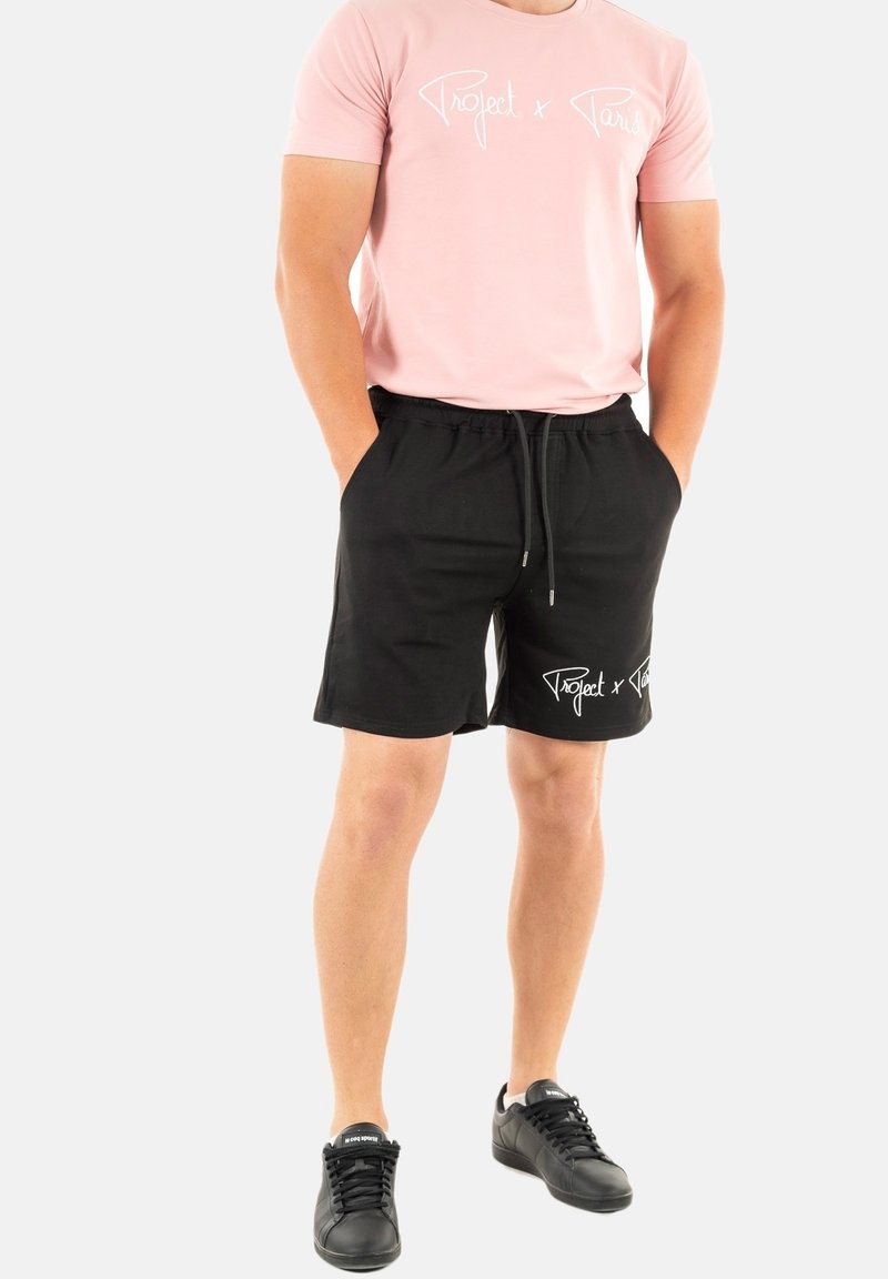Pantalones cortos negros con una cinturilla elástica y cordón, con texto gráfico blanco. Combinados con una camiseta de color rosa claro. Zapatillas negras completan el atuendo.