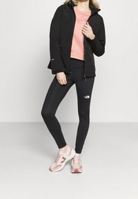Kvinde iført sorte The North Face leggings, lyserød sweatshirt, sort jakke og rosa-hvide sneakers, stående foran en hvid baggrund.