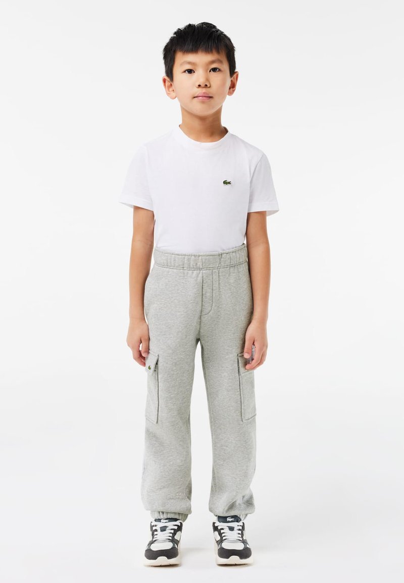 Pantalon de survêtement gris avec des poignets élastiques, des poches latérales et un logo brodé, associé à un t-shirt blanc à manches courtes. Baskets noir et blanc.