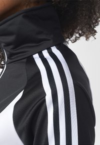 Černá sportovní bunda s vysokým límcem. Na rameni jsou tři bílé pruhy z texturované tkaniny. Celkově hladký černý materiál.